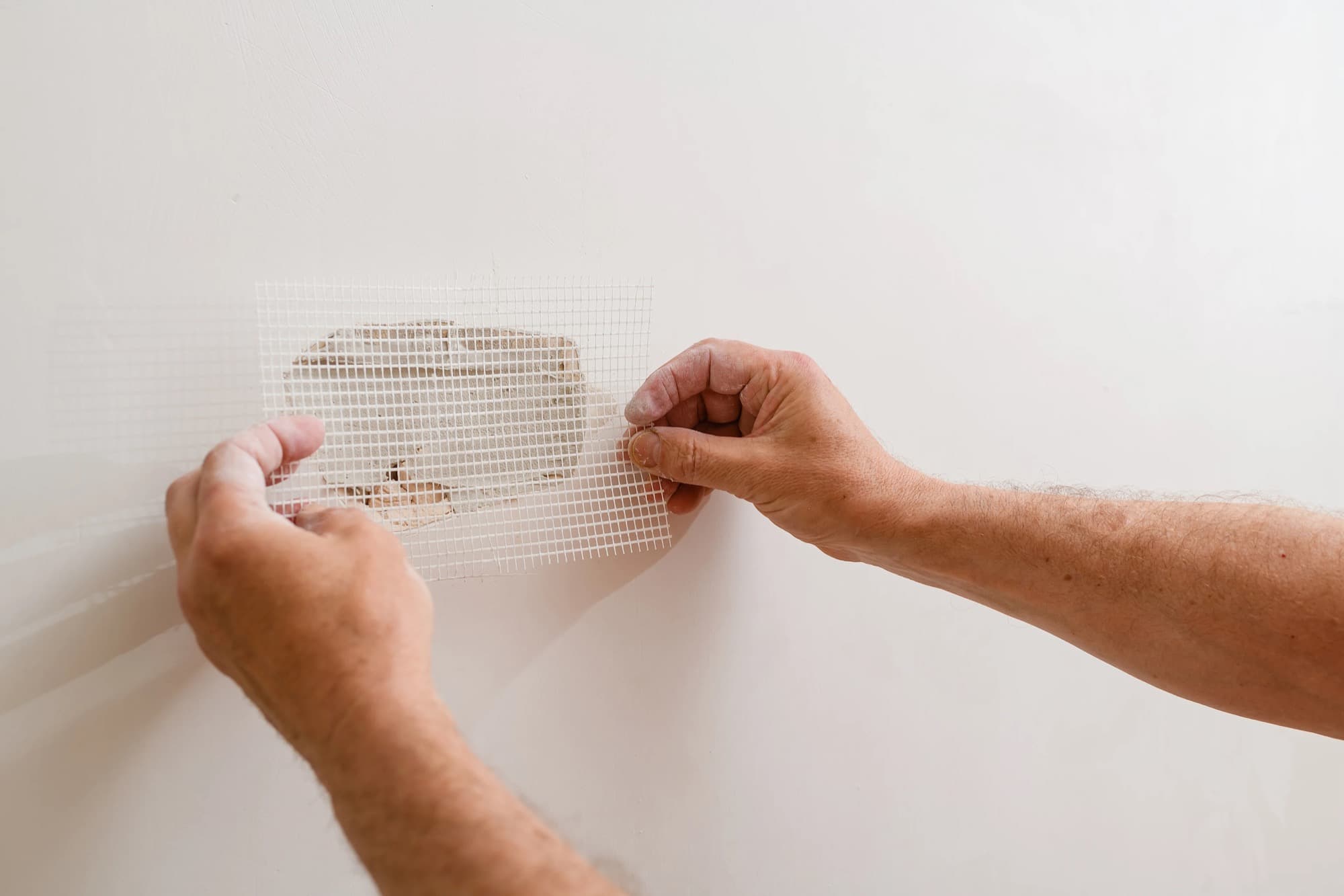 Hands pressing self-adhesive mesh over a patched hole in a plaster wall