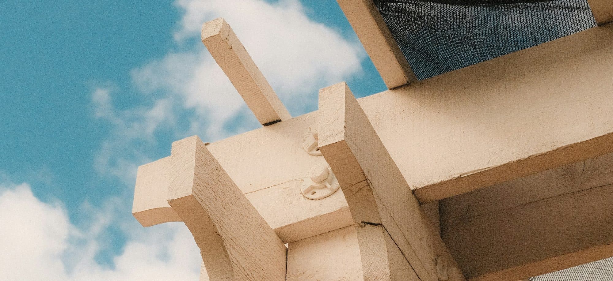 Looking up at a timber pergola beam joint with a shade cloth panel against a blue sky